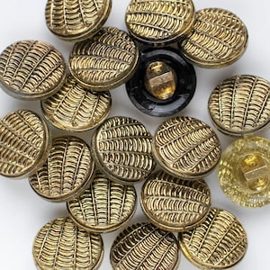May include: A selection of gold-coloured buttons with a textured, ridged design. The buttons are round and have a detailed pattern. One button is black, and another is clear. The buttons are scattered on a white surface.
