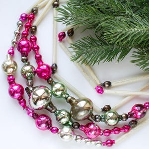 May include: A garland featuring a series of vintage-style glass beads in shades of hot pink, silver, and pale green, interspersed with long, thin, cream-colored straw-like tubes. The decorative strand is arranged in a curved, layered shape.