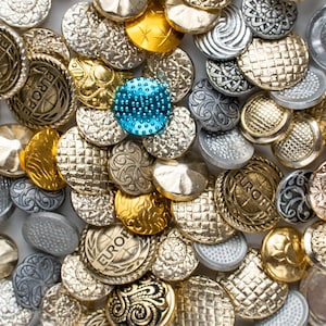 May include: A collection of assorted round metal buttons in gold, silver, and bronze tones with various embossed textures and patterns. One button is bright blue with a textured surface. Several buttons feature the word EUROPE embossed on them.