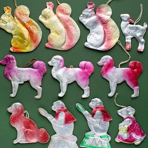 Puede incluir: Una colección de adornos navideños vintage hechos de papel maché. Los adornos tienen forma de animales, incluyendo ardillas, un cordero, perros y un gato. Los adornos están pintados en varios colores, incluyendo rosa, blanco, plateado y amarillo.
