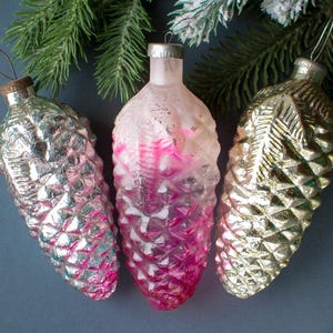 Op de afbeelding: Drie vintage kerstversieringen in de vorm van dennenappels. De ornamenten zijn zilver, goud en roze, met een textuur. Groene dennentakken op de achtergrond creëren een feestelijke vakantiescène.