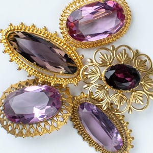 Puede incluir: Un surtido de broches vintage con grandes piedras preciosas facetadas, ovaladas y en forma de marquesa en tonos púrpura y amatista. Los broches tienen engastes de metal dorado ornamentados con intrincados detalles de filigrana y cuentas, sobre fondo blanco.