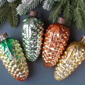 Op de afbeelding: Vier vintage glazen kerstversieringen in de vorm van dennenappels in groen, goud en koper. De ornamenten zijn gerangschikt op een donkergrijze ondergrond met een takje groenblijvend en sneeuw.