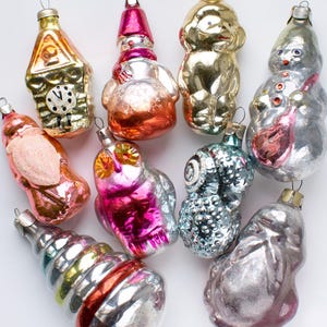 Puede incluir: Una colección de adornos navideños vintage en varias formas, incluyendo una casa, un muñeco de nieve, un búho y un perro. Los adornos son de vidrio y presentan colores metálicos dorados, plateados, rosas y azules. Cada adorno tiene un lazo metálico para colgar.