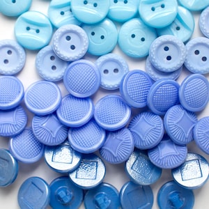 May include: A collection of round buttons in various shades of blue, ranging from light sky blue to deep periwinkle. The buttons feature diverse textures, including smooth surfaces with two holes, raised dot patterns, starburst designs, and square centres.
