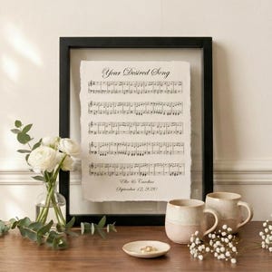 May include: A framed print of sheet music with the text "Your Desired Song." The black framed print is displayed on a wooden table, alongside a small vase of white flowers, two mugs, and a small dish with rings.