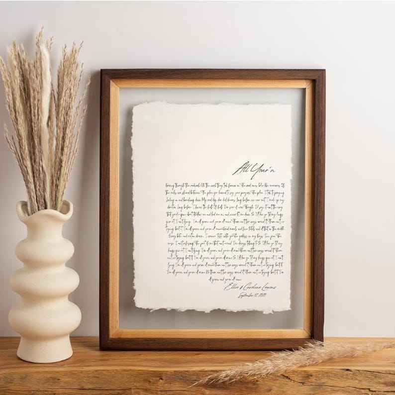 May include: A framed artwork featuring handwritten text on textured paper. The frame is a combination of dark and light wood tones. The artwork is displayed with a vase of dried plants on a wooden surface.