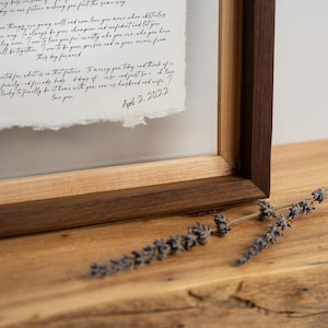 May include: A framed document featuring handwritten black cursive text on textured white paper with deckled edges. The frame is constructed from dark wood with a light wood inner border. The text includes the date April 2, 2022.