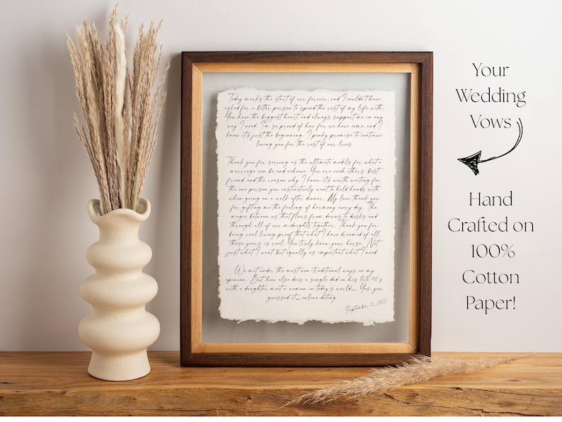 May include: A rectangular dark wood floating frame containing a piece of off-white paper with handwritten black cursive text. The paper has rough, deckled edges. To the left is a matte cream-coloured vase filled with dried beige pampas grass.