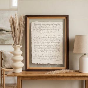 May include: A rectangular floating frame with a dark wood border containing a piece of off-white handmade paper with deckled edges. The paper features handwritten black cursive text detailing personal vows dated September 21, 2024.