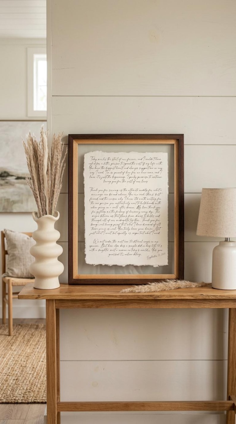 May include: A rectangular floating frame with a dark wood border containing a piece of off-white handmade paper with deckled edges. The paper features handwritten black cursive text detailing personal vows dated September 21, 2024.