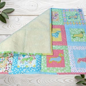 May include: A patchwork quilt with dachshund dog designs on squares of blue, green, pink, and yellow. The quilt has a cream-coloured fleece backing. The squares are arranged in a grid pattern.