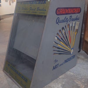 May include: A vintage metal display case with a glass front showcasing M. Grumbacher paint brushes. The case is painted gray and features the text "M. Grumbacher Inc. The World's Finest Brushes" and "Grumbacher Quality Brushes For Art and Industry".