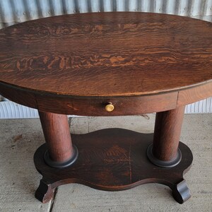May include: Oval-shaped antique wooden table with a dark brown finish. The table has a drawer with a brass knob, supported by two cylindrical legs and a lower shelf. The table is approximately 30 inches in diameter.