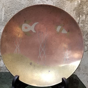 May include: A round, decorative metal plate with a copper and brass finish. The plate features etched designs of fish and seaweed. The plate is displayed on a black metal stand, showcasing its decorative appeal.