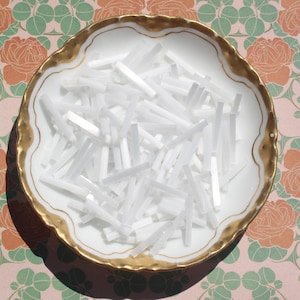Peut inclure: Un plat en porcelaine blanc avec un bord doré rempli de nombreux petits objets rectangulaires blancs. Le plat repose sur un fond rose avec un motif floral de roses oranges et de feuilles vertes.