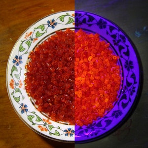 May include: A white ceramic plate with a floral design, filled with small, orange-red beads. The beads glow brightly under a blacklight, revealing a fluorescent orange color.