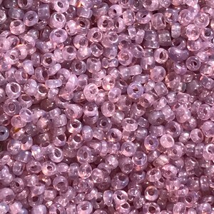 May include: A close-up view of a large quantity of small, translucent, lavender-colored glass beads. The beads are round with a hole in the center.