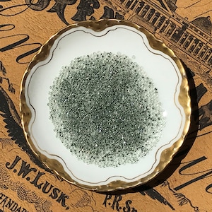 Puede incluir: Un pequeño plato de porcelana blanca con un borde dorado, lleno de una gran cantidad de pequeñas cuentas de vidrio verde transparentes.
