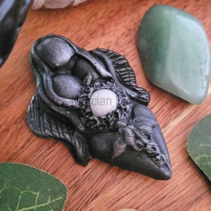May include: A dark gray, sculpted pendant with a central, light-colored orb. The pendant has intricate details and is surrounded by green stones and a leaf on a wooden surface. The word "clan" is visible on the orb.