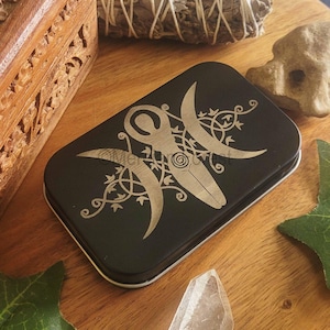 May include: A black metal tin with a silver design of a triple moon symbol and a stylized female figure. The design is surrounded by vines.