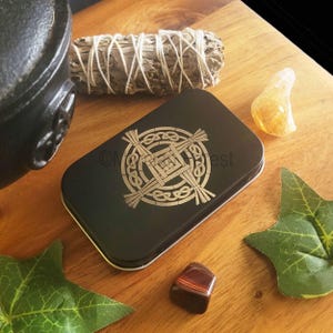 May include: A black metal tin with a gold Celtic cross design. The tin is sitting on a wooden surface with a sage smudge stick, a yellow crystal, and a red tiger's eye stone.