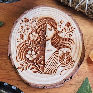 May include: A wooden round disc with a burned-in design of a woman with flowers and a pomegranate. The woman's face is split in half, with one side showing a more natural look and the other side showing a more stylized look.