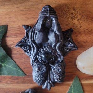 May include: A dark, detailed sculpture of a female form with a headdress, flowing hair, and owl-like elements. The sculpture has a textured finish and is placed on a wooden surface with green leaves and a white stone.