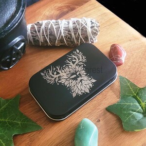 May include: A black rectangular tin featuring a silver tree design and the word "forest". It is surrounded by a sage bundle, crystals, and green leaves on a wooden surface.