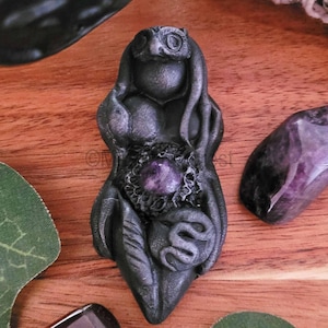 May include: A dark gray pendant with an owl-like face, a central purple gemstone, and a snake design. The pendant has a carved, textured appearance and is likely made of a stone or resin material. The overall design suggests a mystical or spiritual theme.