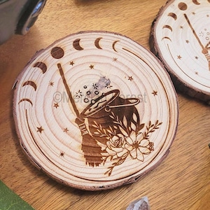 May include: A wooden coaster with a laser-etched design of a cauldron, broom, and moon phases. The design is in black and white and features a floral motif.
