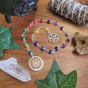May include: A rainbow colored crystal pendulum with a wooden pentagram charm and a silver pentagram charm. The pendulum is hanging from a silver chain. The pendulum is on a wooden surface with green leaves and a white crystal.