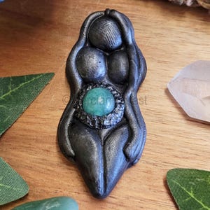 May include: A black ceramic figurine of a woman with a green gemstone in her belly. The figurine is a representation of the female form and is often used in spiritual practices.