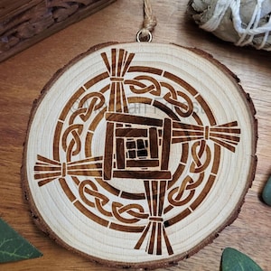 May include: Circular wooden ornament featuring a Celtic knot design. The ornament is crafted from a cross-section of a tree branch, with the design etched into the light-coloured wood. A twine loop is attached for hanging. The design includes a central square and radiating elements.