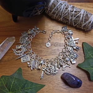 May include: A silver charm bracelet featuring a range of charms, including a crescent moon, a star, a pentagram, a sun, a tarot card, an owl, a tree, a leaf, a goblet, a wand, a cauldron, a crystal, and an acorn. The bracelet is displayed on a wooden surface with a green leaf and a purple crystal.