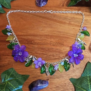May include: A silver chain necklace with purple and green flower charms, silver fairy charms, and green leaves. The necklace is on a wooden surface.
