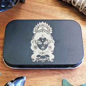 May include: A black rectangular tin featuring a silver-coloured image of a deity with a crown, earrings, and a necklace. The deity has a long tongue and is surrounded by skulls. The tin has a silver-coloured rim.