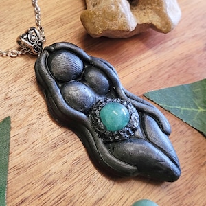 May include: A black polymer clay pendant necklace featuring a stylized female figure with three round shapes and a green gemstone in a textured setting.