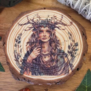May include: A round wooden coaster with a printed image of a woman adorned with a crown of antlers and foliage. The artwork is set on a natural wood slice, displaying the tree's growth rings. The woman's clothing is in shades of green and brown, creating an earthy aesthetic.