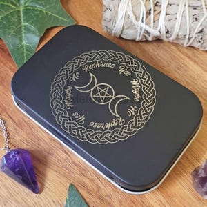 May include: A black rectangular tin with a silver Celtic knot design, crescent moons, and a pentagram. The text on the tin reads "Yes, Maybe, No, Rephrase". A purple crystal pendulum and sage bundle are also visible.