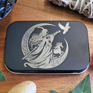 Peut inclure: Une boîte en métal noire avec un motif gravé argenté. Le motif représente une femme aux longs cheveux, un croissant de lune, un cheval et un oiseau. La boîte a un bord argenté et est de forme rectangulaire.