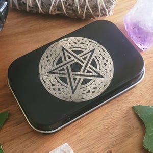 May include: A black metal tin featuring a silver pentagram design. The pentagram is encircled by a Celtic knot design.