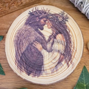 May include: A wooden coaster with a printed image of a couple embracing. The man is wearing a crown of antlers and a fur-lined cloak. The woman is wearing a floral crown and a white dress. The image is in shades of purple, brown, and white.