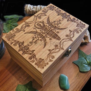 May include: A wooden box with a hinged lid, featuring an intricate laser-engraved design of a triple moon symbol surrounded by floral vines. The box is made of light brown wood and has a brass latch.
