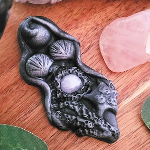 May include: A dark grey pendant with a central light purple stone, surrounded by intricate, textured details. The pendant features shell-like designs and organic shapes. The background includes a pink crystal and a wooden surface.