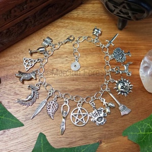 May include: Silver charm bracelet featuring a range of charms, such as a pentacle, a crescent moon, a feather, a leaf, a cauldron, a sun, a tree, a sword, a broom, a hand holding a wand, a goblet, a bird, a book, and an acorn.