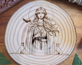 May include: A wood-burned design on a round wooden coaster features a woman with long flowing hair, wearing a cloak and holding a feather. Two cats flank her on either side.