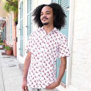 May include: A white short-sleeved button-down shirt with a red lobster print. The shirt is worn by a person with curly hair.
