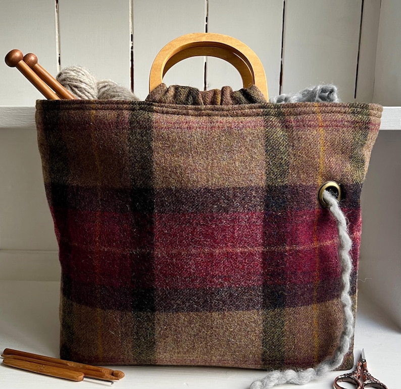 May include: A brown, burgundy, and green plaid fabric project bag with rounded wooden handles. The bag features a metal grommet on the front for yarn to pass through, and it is filled with gray yarn and wooden knitting needles.