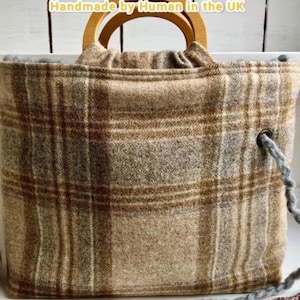 May include: A tote bag featuring a brown, tan, and gray plaid pattern with rounded wooden handles. The bag includes a gray yarn drawstring threaded through a metal eyelet on the side. The text "Handmade by Human in the UK" is visible at the top.